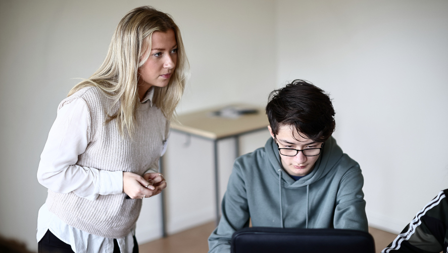 Lärare står till vänster om elev som sitter framför dator.