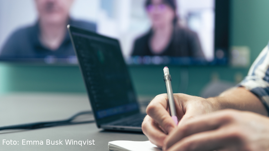 Person antecknar i förgrund, med ett digitalt möte på skärm i bakgrunden.