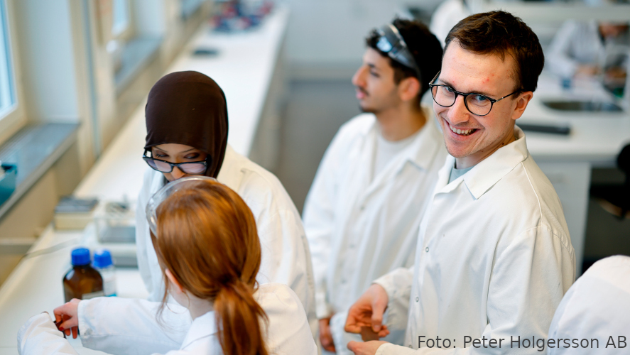 Lärare med tre elever vid en labbänk, alla med vita labbrockar.