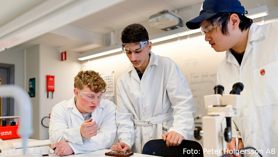 Tre elever med labbrockar fokuserar på något på en bänk.